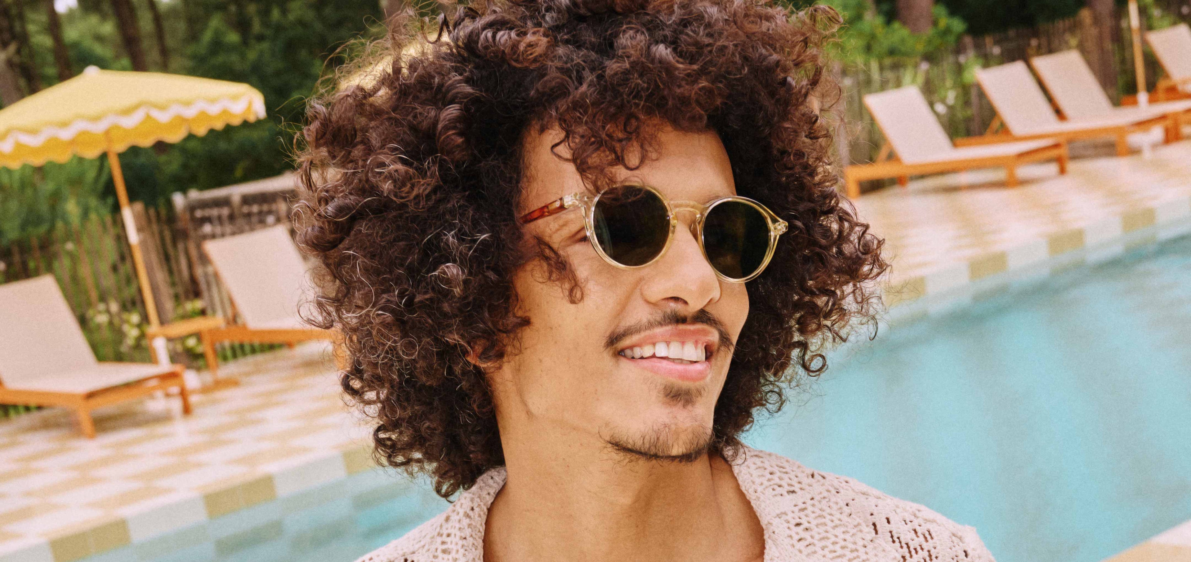 A smiling man with voluminous curly dark brown hair and a light goatee, wearing clear-framed round sunglasses and an off-white crocheted knit shirt, looks to the right against a background of a swimming pool, lounge chairs, and a yellow and white fringed umbrella.